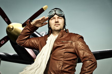 Portrait Of A Vintage Pilot With Leather Cap, Scarf And Aviator Glasses Salutes In Front Of A Historic Airplane - Portrait Of A Man In Historical Pilot Clothing