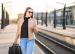 © terovesalainen - Woman walking in train station and using smartphone. Passenger buying electronic digital ticket with mobile phone. Happy lady pulling suitcase and luggage in platform.