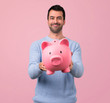 Man with blue sweater holding a big piggybank on pink background