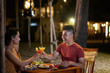© DragonImages - Affectionate young couple clinking glasses of alcoholic cocktails together while enjoying warm evening at beach restaurant of tropical resort