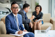 © DragonImages - Portrait shot of handsome Asian car dealer wearing elegant suit looking at camera with wide smile while distracted from working meeting at showroom