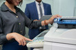© DragonImages - Close-up shot of smiling office assistant using multi-function printer in order to make copy of document, male colleague with digital tablet in hands standing behind her
