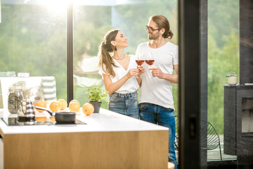 Young couple standing together with wine near the window of the modern apartment with green area outdoors celebrating housewarming