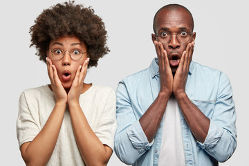 Wall Mural - Lovely African American couple feels puzzled and embarrassed, have scared looks at camera, see fire of neighbour`s house, stare at camera with widely opened eyes and mouth, stand against white wall