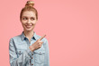 © wayhome.studio  - Adorable young woman with hair bun, has positive expression, indicates at blank copy space, wears denim jacket, stands alone against pink background. Delighted female shop assistant advertises clothes