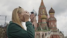 Side View Of A Blonde Young Tourist Wearing A Green Coat Making Photos Of Saint Basil's Cathedral. Moscow, Russia. Cloudy Day. Blurred Background. Handheld Real Time Medium Shot.