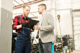 Fototapeta  - Young bearded auto mechanic looking at checklist and smiling while male customer thanking him for maintenance in auto repair service