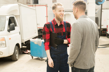 Young bearded male auto technician in workwear standing in repair shop, talking to his acquaintance and smiling happily