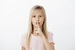 © Cookie Studio - Studio shot of sctrict confident little blond girl in casual pink t-shirt, saying shh while showing shush gesture with index finger over mouth, asking to be quiet while looking after baby brother