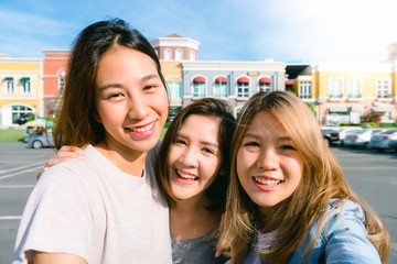 Wall Mural - Close up of young Asian women group selfie themselves in the pastel buildings city in nice sky morning. City lifestyle of young women group on weekend. Outdoor shopping and city lifestyle concept.