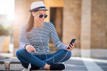 Young woman sitting on a city street  and drinking coffee to go and using mobile phone