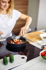 Blonde caucasian girl preparing meat kebabs, frying at non-stick pan on the stove, the process of cooking kebab at home interior