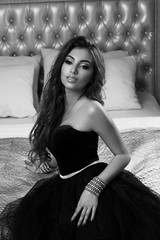 Wall Mural - Young beautiful tanned woman with long wavy hair in luxurious black evening dress sitting at bed in luxury interior