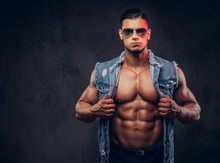 Sexy Naked Fashionable Athletic Man In A Denim Vest And Sunglasses Poses In A Studio.