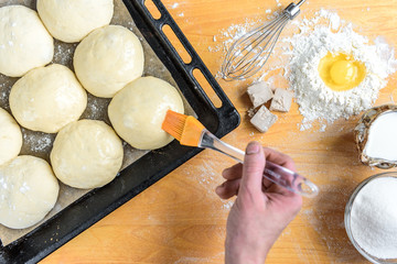 Making sweet bread dough top view. Women's hands lubricate the buns with egg. Cooking ingredients for pastry on rustic wood sprinkled with flour, sugar, milk and yeast.