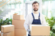 © pressmaster - Serious confident handsome bearded moving company employee in overalls carrying heavy cardboard box and looking at camera in lobby, stack of boxes behind him