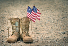 Old Military Combat Boots With Dog Tags And Two Small American Flags. Rocky Gravel Background With Copy Space. Memorial Day Or Veterans Day Concept.
