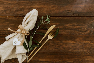 Wall Mural - Gold cutlery with linen napkin and olive branch on wooden background. Wedding festive tableware