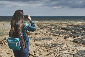 Wall Mural - Back view of cheerful young woman photographing beautiful seascape while standing on rocky shore. She is using her smartphone. Copy space 