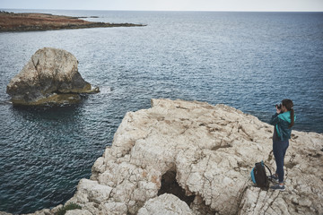 Wall Mural - Calm young woman is taking photos of beautiful sea landscape. She is standing on rock mountain near her backpack and holding camera 