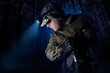 Profile view photo of a male person in brown tactical outfit jacket, gloves, backpack and head flashlight taking out knife on night woods background.