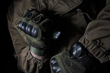 Photo of a male person in brown tactical outfit jacket and gloves using green tactical led flashlight and military watch.