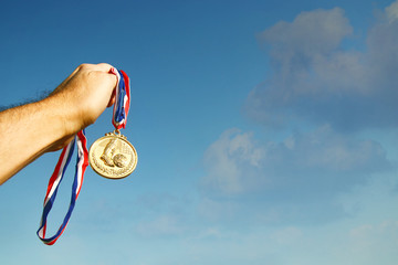 Wall Mural - man hand raised, holding gold medal against sky. award and victory concept.