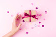 Woman hand and kraft gift box with pink bow on pink background decorated with confetti.. Top view, holiday present concept.