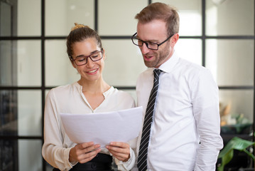 Wall Mural - Smiling coworkers talking and discussing document. Business man and woman standing in office with glass wall in background. Business result concept.