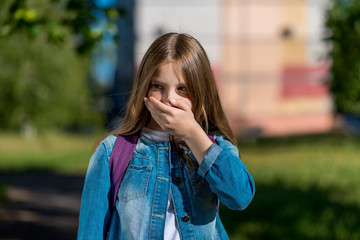 A little blond girl covered her mouth with her hand. Emotions of fear of fear. Summer outdoors in fresh air. The teenager looks away in surprise. At the break after school.