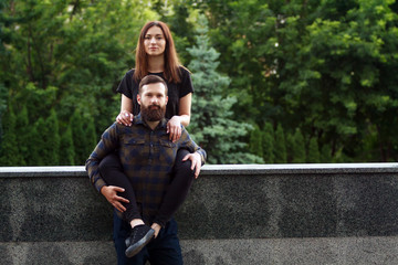 Young couple sitting on parapet in park. Attractive woman and handsome bearded man are standing on street in the city.