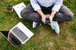 Cropped photo from top of caucasian man in business clothing, sitting on grass in park while using cell phone and silver laptop