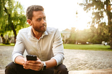 Image Of Handsome Guy In White Shirt, Sitting In Park And Looking Aside While Holding In Hands Black Cell Phone
