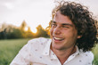 Close up portrait of happy young male with curly hair, smiling and relaxing on the green lawn in the park, listening funny story. Copy space for advertising. People and lifestyle concept.