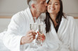 Image closeup of caucasian handsome man kissing beautiful asian woman on cheek with closed eyes, while clinking glasses with champagne at hotel room