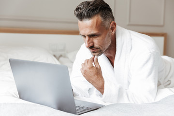 Sticker - Photo of european man with gray beard wearing white bathrobe using silver laptop, while lying on bed in bedroom or hotel apartment