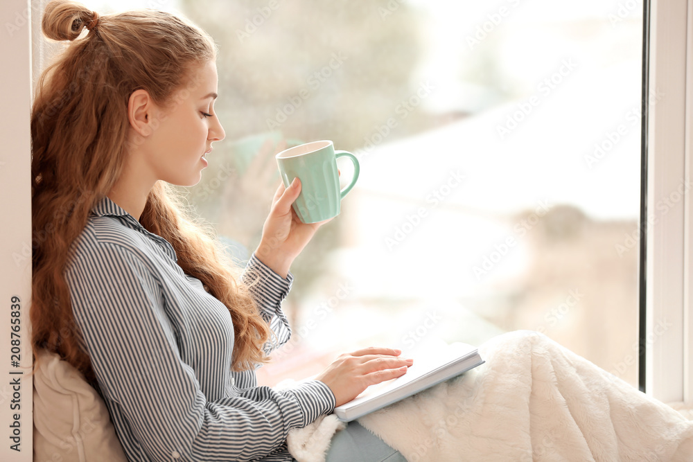 Young woman drinking tea while reading book near window indoors