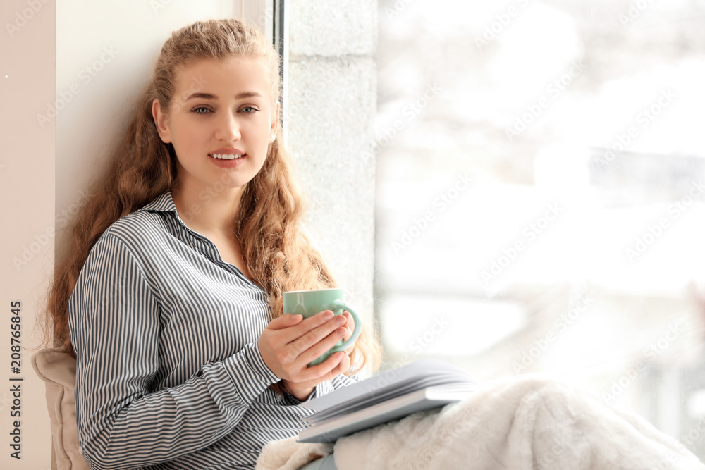 Young woman drinking tea while reading book near window indoors