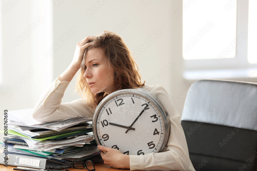 Mature tired woman with clock at table in office. Time management concept