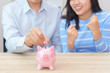 © petzshadow - Smile couple putting a coin into a pink piggy bank on wooden desk - save money for the future.