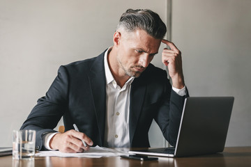 Canvas Print - Concentrated thoughtful business man 30s wearing white shirt and black suit working at laptop in office, while writing down notes