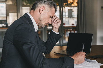 Canvas Print - Image of handsome caucasian businessman wearing white shirt and black suit sitting at table in office, during work with documents and laptop