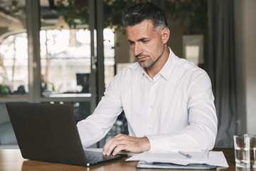 Sticker - Image of concentrated confident businessman 30s wearing white shirt sitting at table in office, during work with documents and laptop