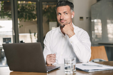 Wall Mural - image of european businessman 30s wearing white shirt sitting at table in office, and working at lap