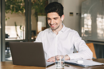 Poster - Image of handsome businessman wearing white shirt and modern earpod smiling while sitting at table in office, and looking at laptop