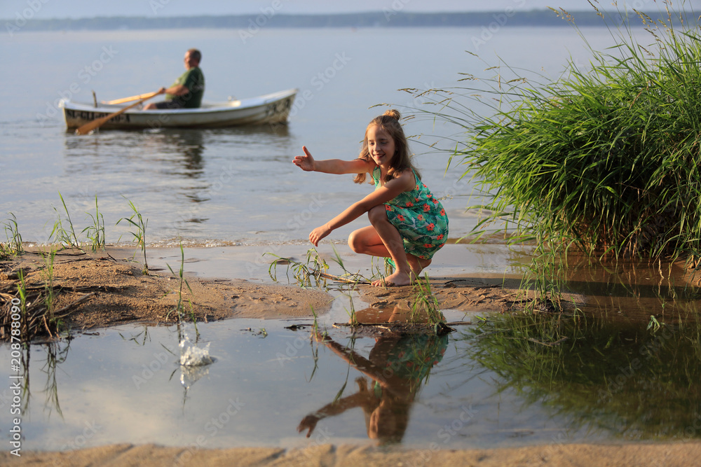 Uśmiechnięta dziewczynka wrzuca kamienie do wody jeziora, mężczyzna na łodzi. - obrazy, fototapety, plakaty 