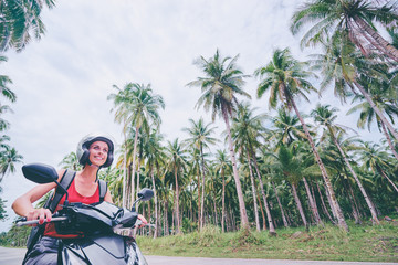 Wall Mural - Tropical travel and transport. Young beautiful woman in helmet riding scooter on the road with palm trees.