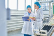 © Seventyfour - Portrait of  two young female workers wearing lab coats and hair covers standing by  power units and writing on clipboard  in clean production workshop, copy space