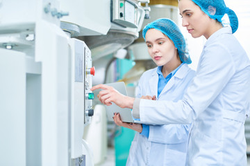 Wall Mural - Side view portrait of  two young female workers wearing lab coats setting up power units and pushing buttons on control panel in clean production workshop, copy space