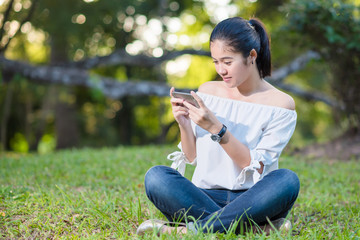 Beautiful Young asian woman smiling while reading her smartphone in garden park outdoor.